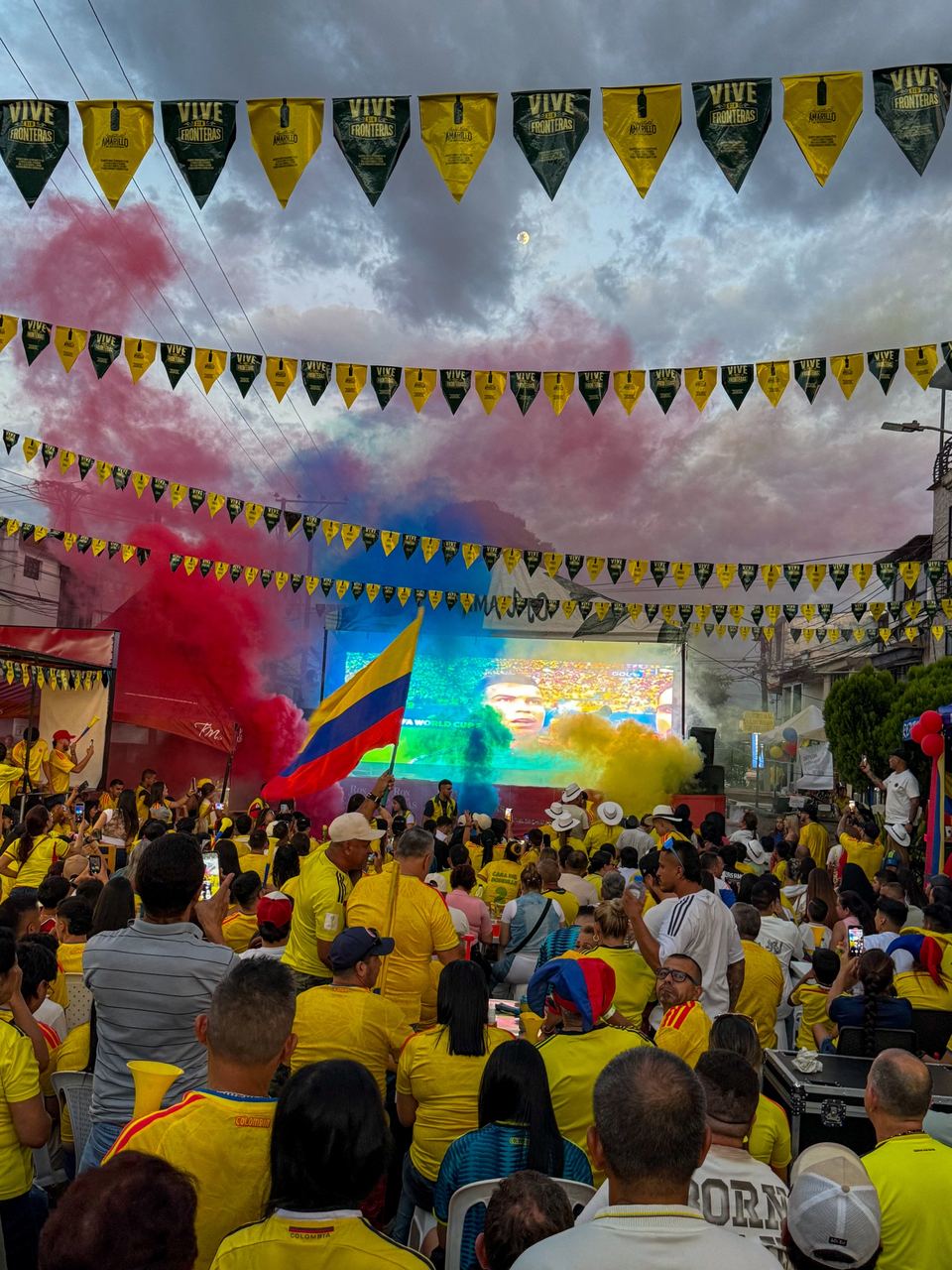 Multitud en la vía pública viendo una transmisión en una pantalla LED gigante, con humo de colores